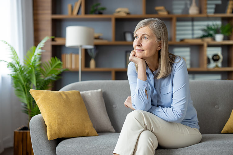 A thoughtful senior woman sits alone on a couch, looking away with a reflective expression